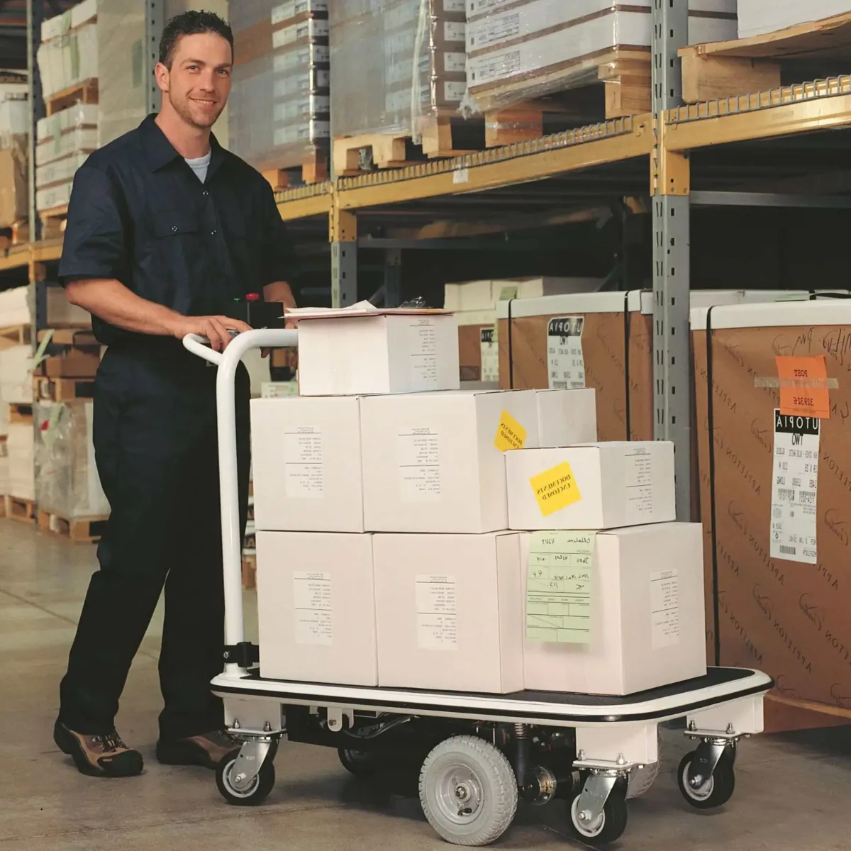 Operator driving a Pony Express 1031 motorized platform cart loaded with boxes in a warehouse aisle
