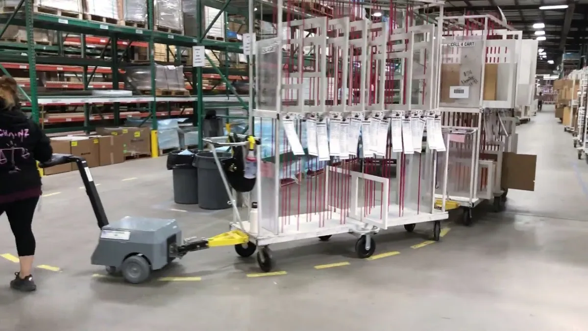 Operator walking ahead of an electric tugger that tows a long train of tall carts down a warehouse aisle